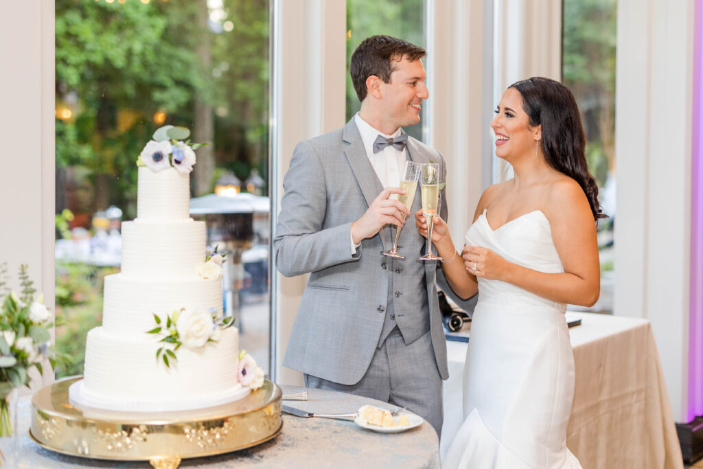 bride and groom cheers with champagne glasses next to wedding cake