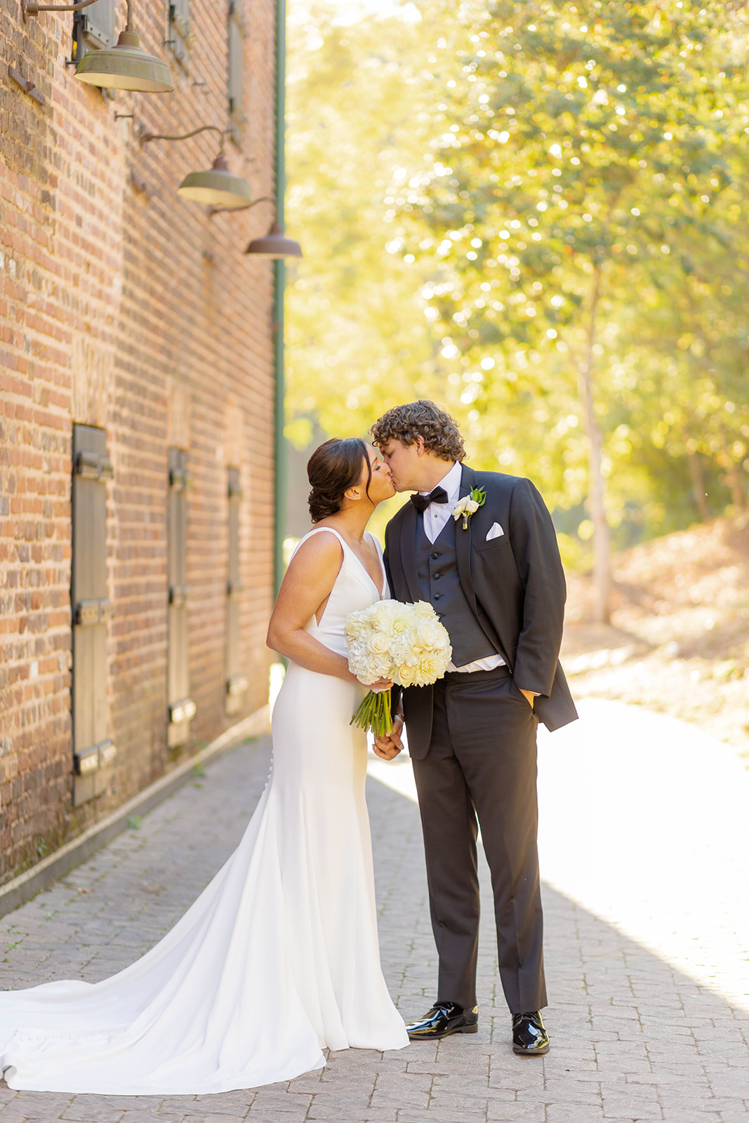 Newlyweds kiss and hold hands on a brick paved path behind the building at sunset