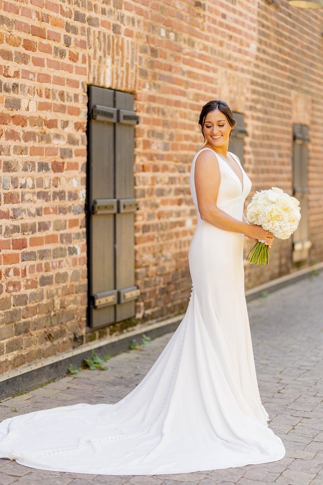 A bride smiles in her silk dress while looking down her shoulder and holding her bouquet in an alley