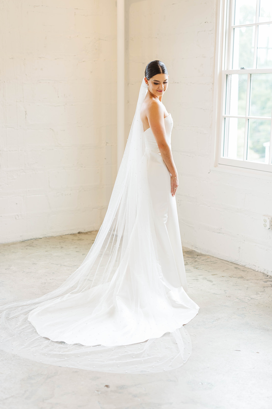 A bride smiles down her shoulder while admiring her gown in a window