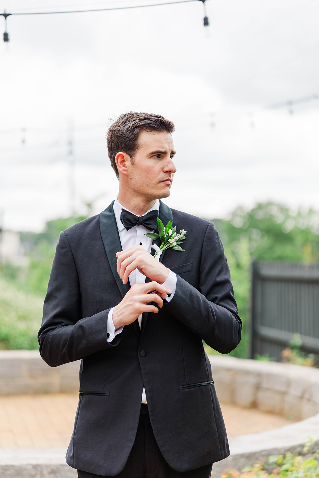 A groom looks over his shoulder while adjusting his cufflinks in a garden