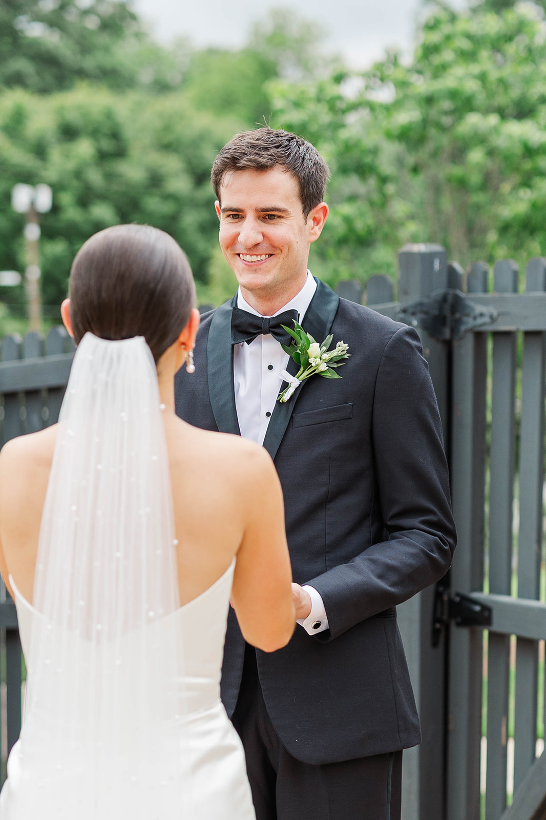 A groom in a black tux smiles big during his outdoor first look