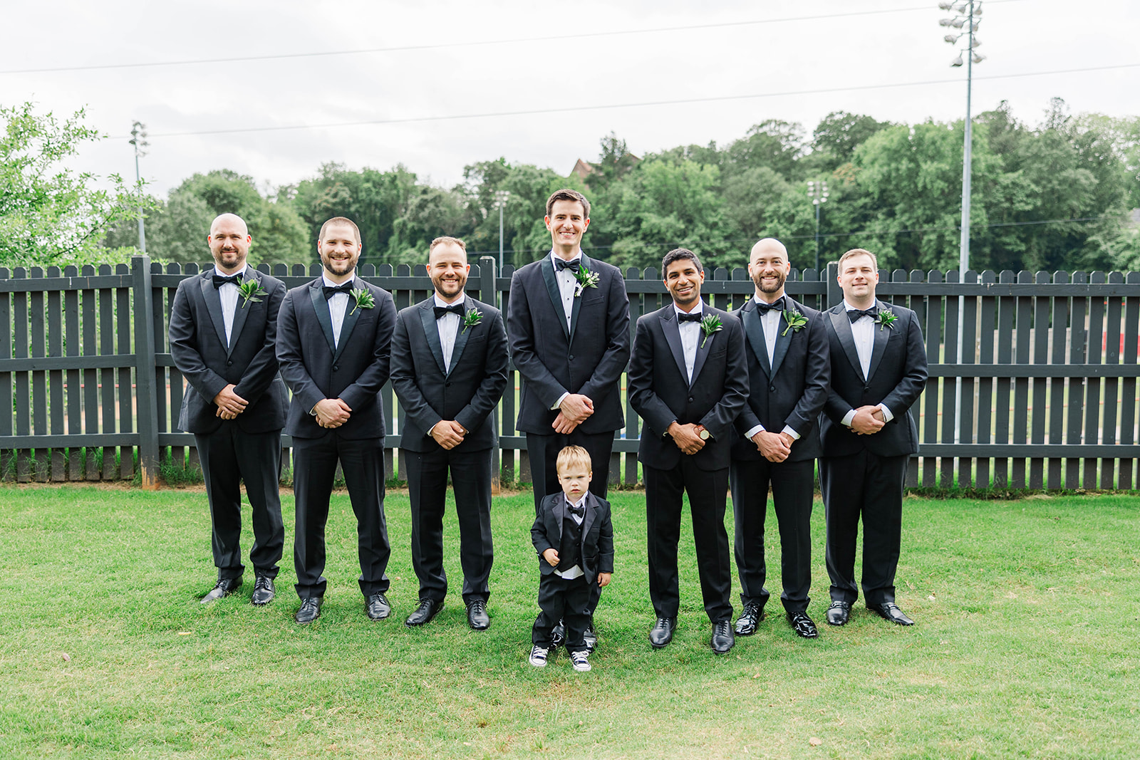 A groom stands in a lawn in black tuxedo with his groomsmen and toddler ring bearer