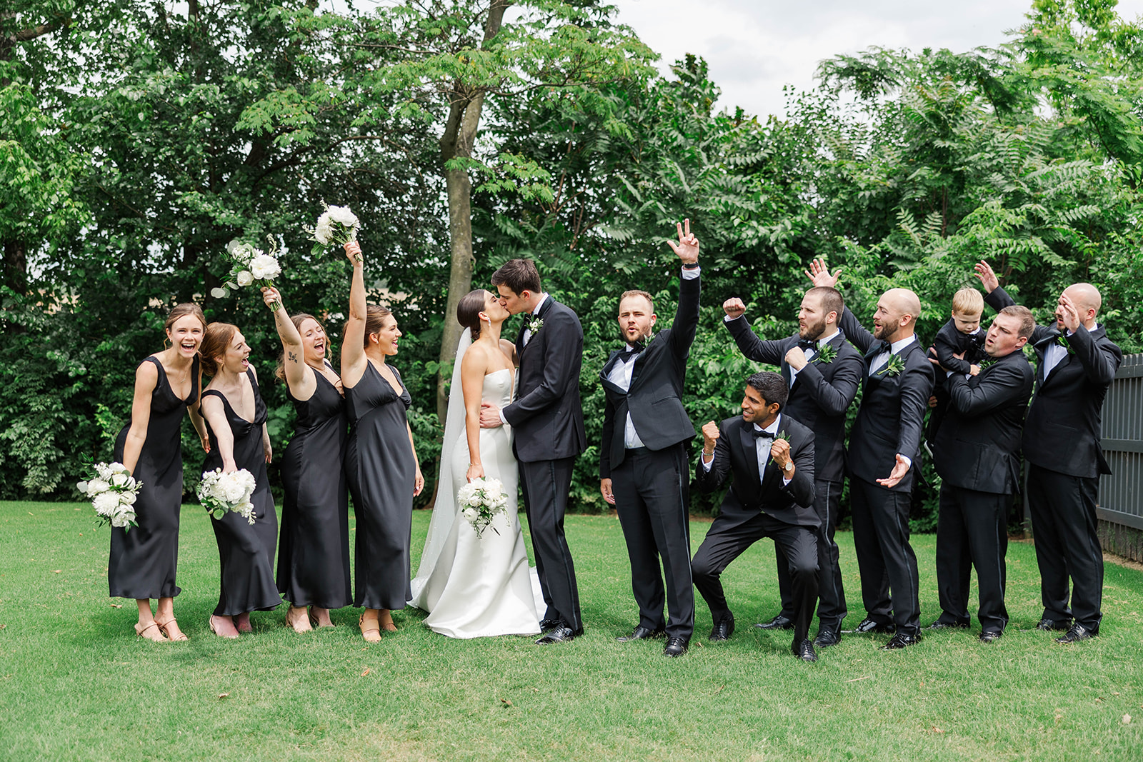 Newlyweds kiss in the lawn as their wedding party cheers and celebrates