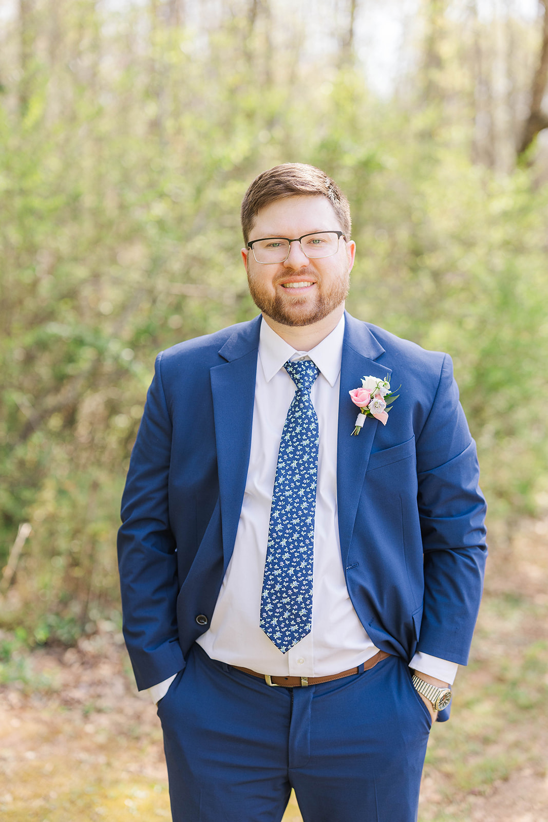 A happy groom smiles in a blue suit with hands in his pockets outside