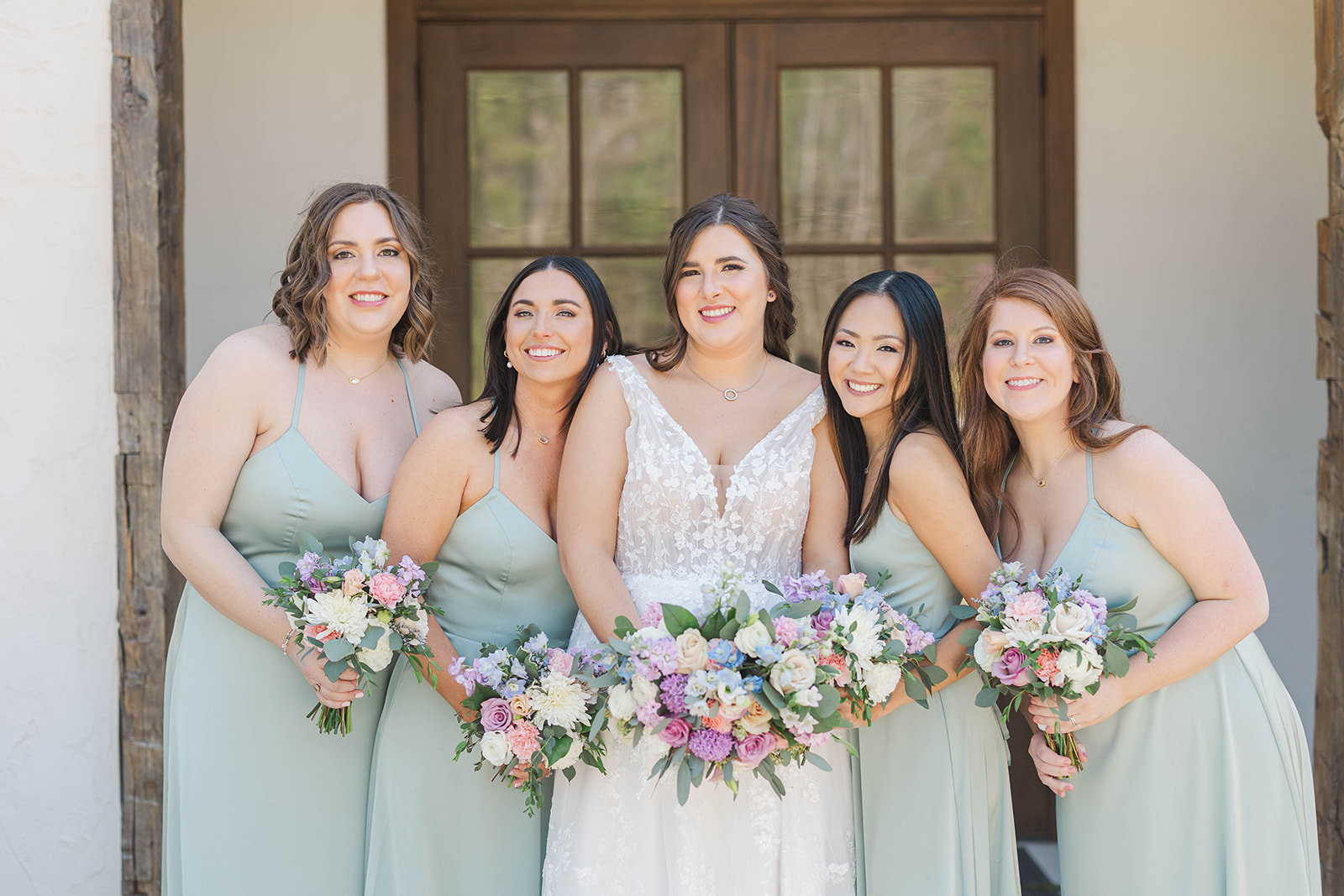 A bride smiles in her lace dress while holding bouquets with her four bridesmaids at White Fox Cottage