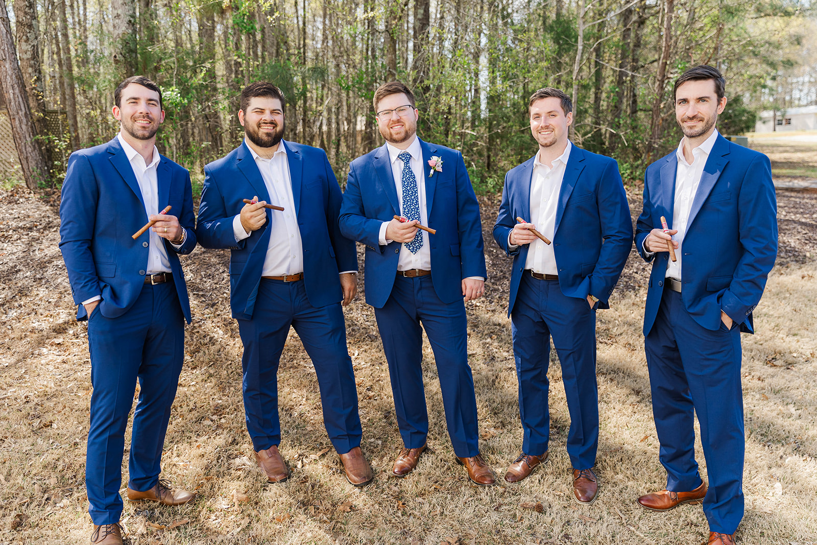 Groomsmen in blue suits stand in the lawn by the forest holding cigars