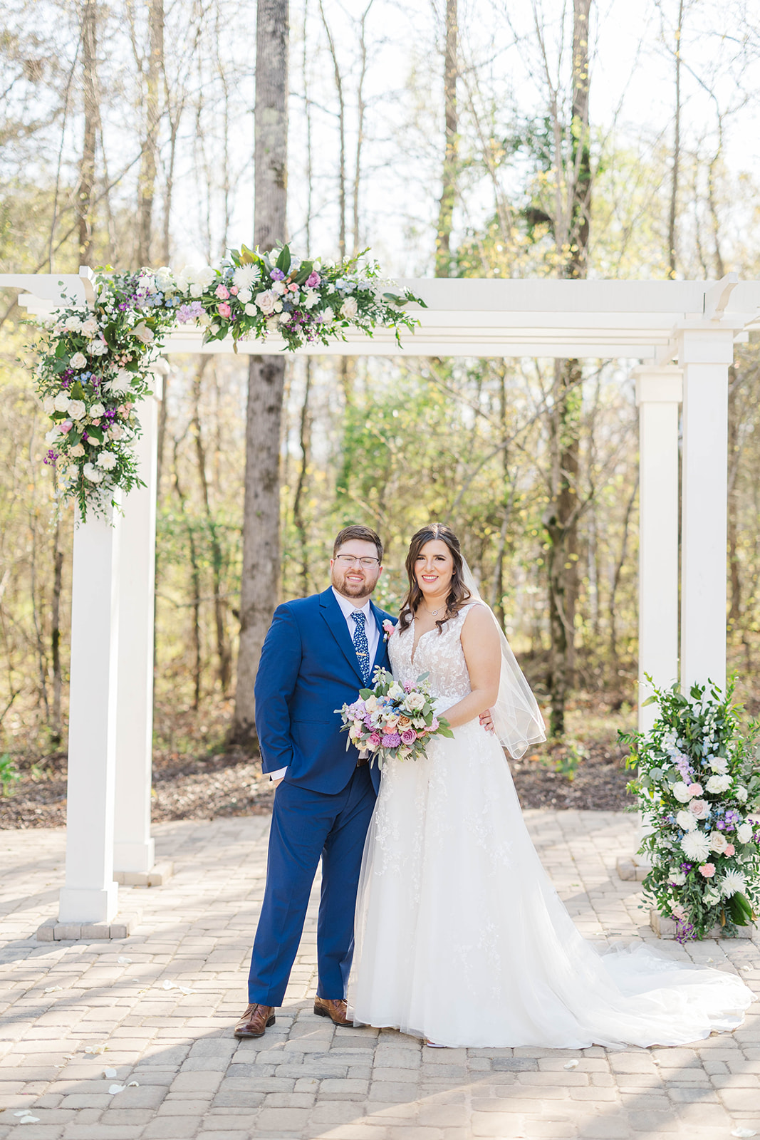 A bride and groom smile while standing together under the flower covered white pergola