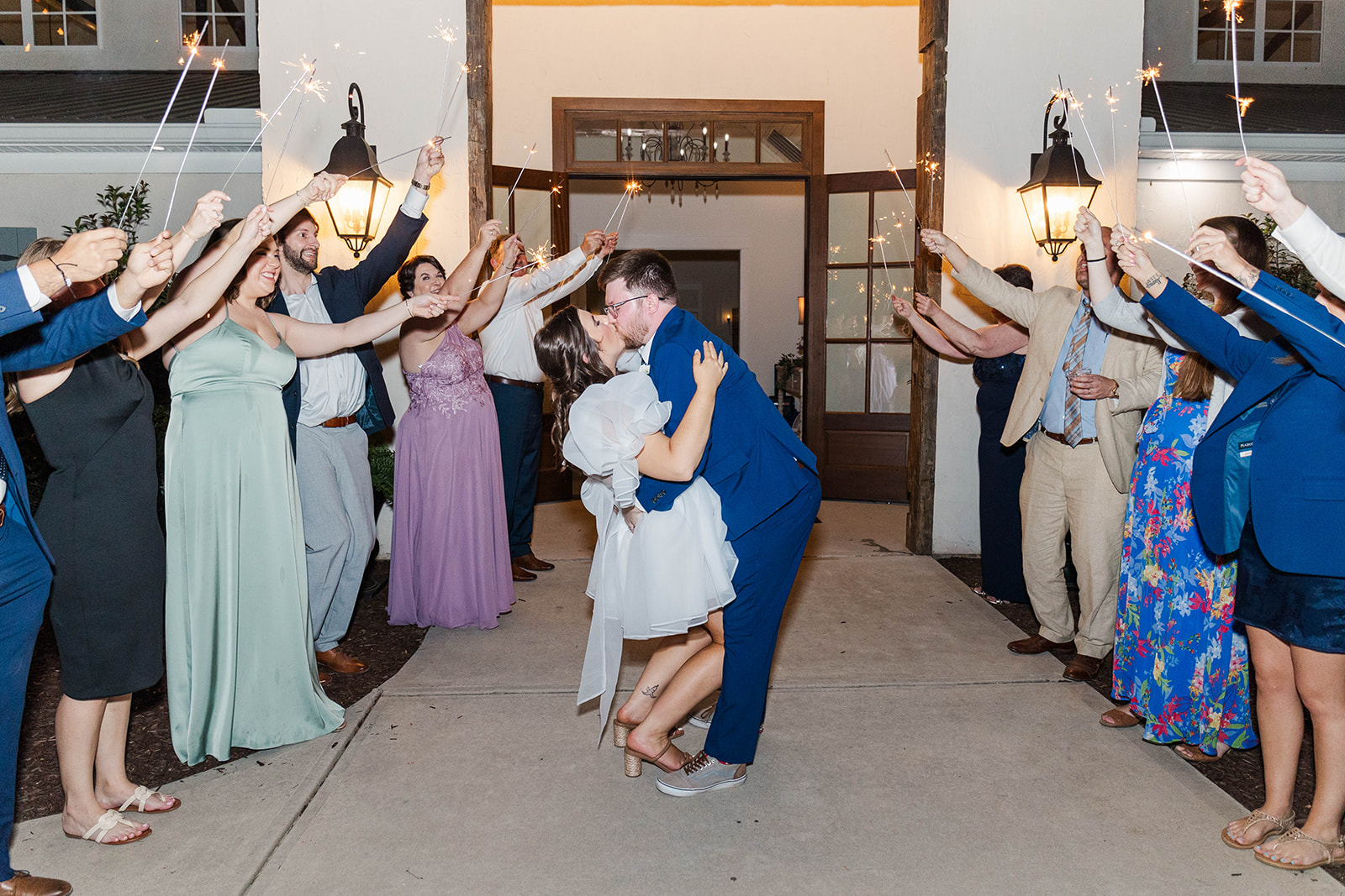 Newlyweds kiss while celebrating under sparklers held by guests while leaving White Fox Cottage