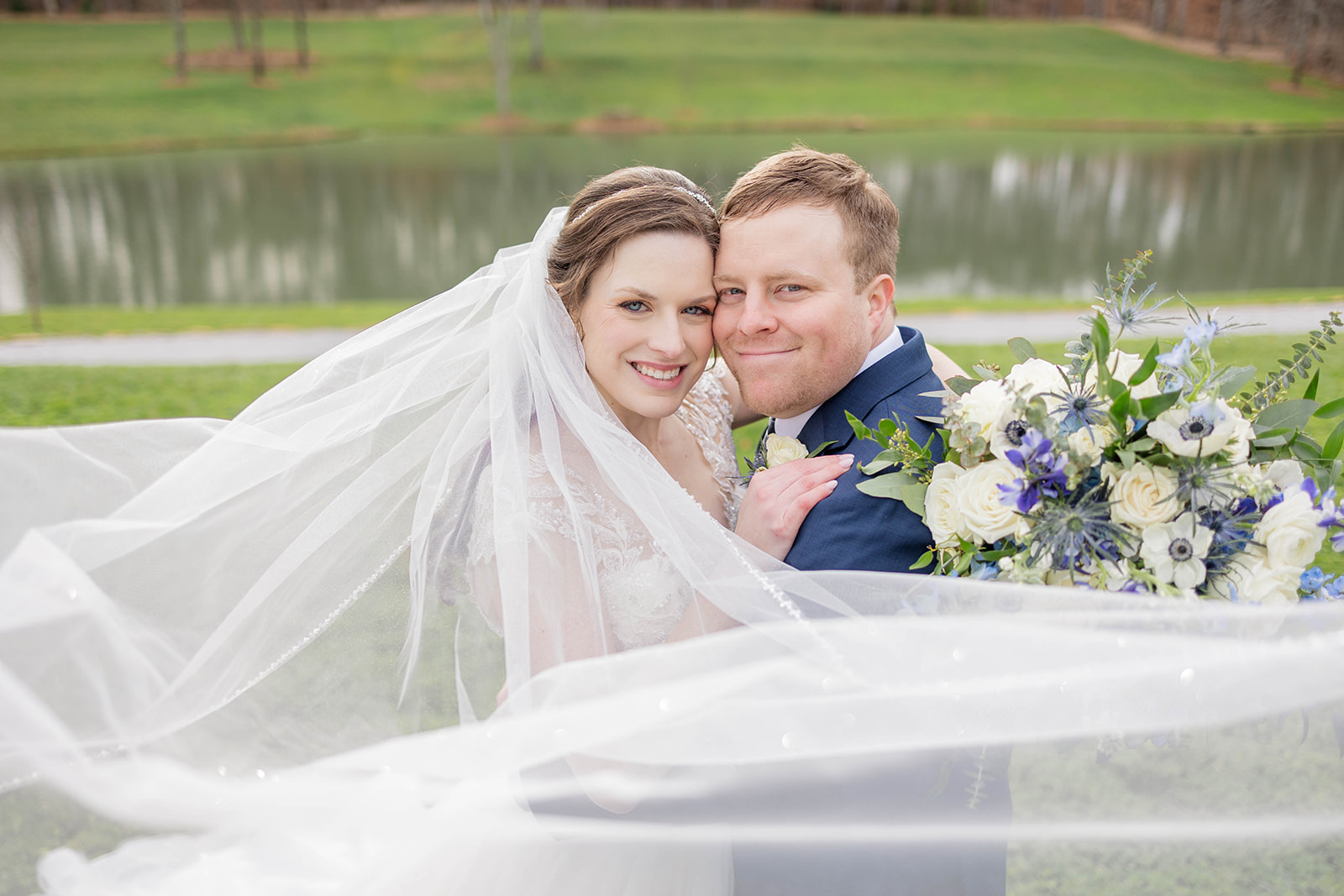 A bride and groom touch cheeks while standing in a lawn by a pond wrapped in the veil