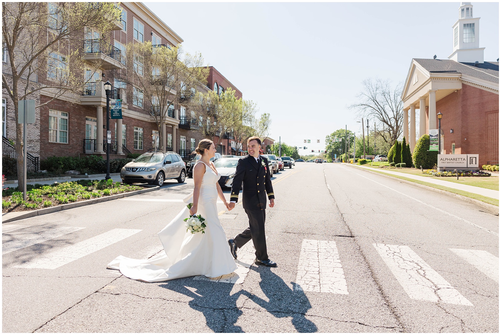 Newlyweds smile at each other while holding hands and walking across a crosswalk to the church