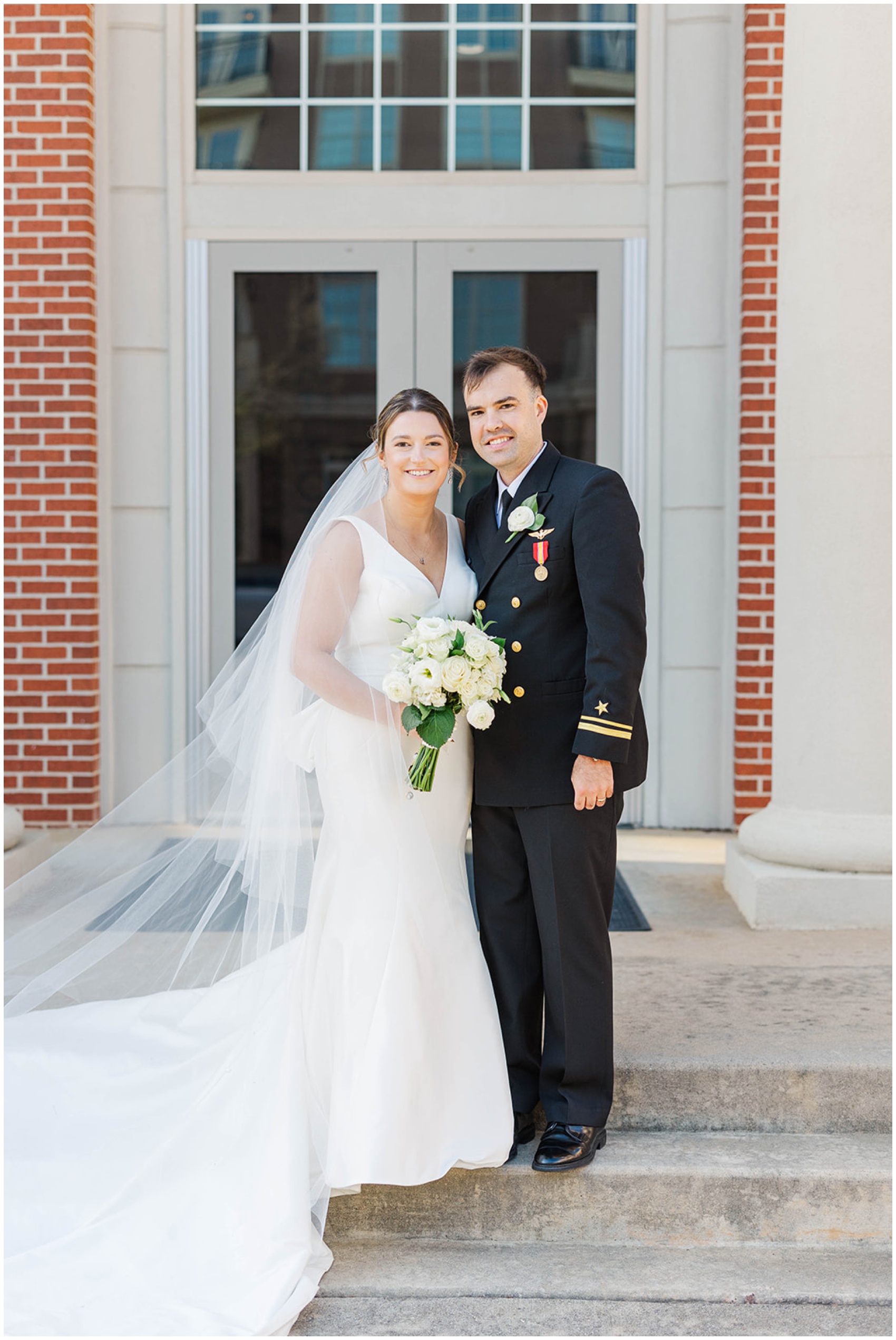 A bride and groom stand at the steps of the chapel smiling together
