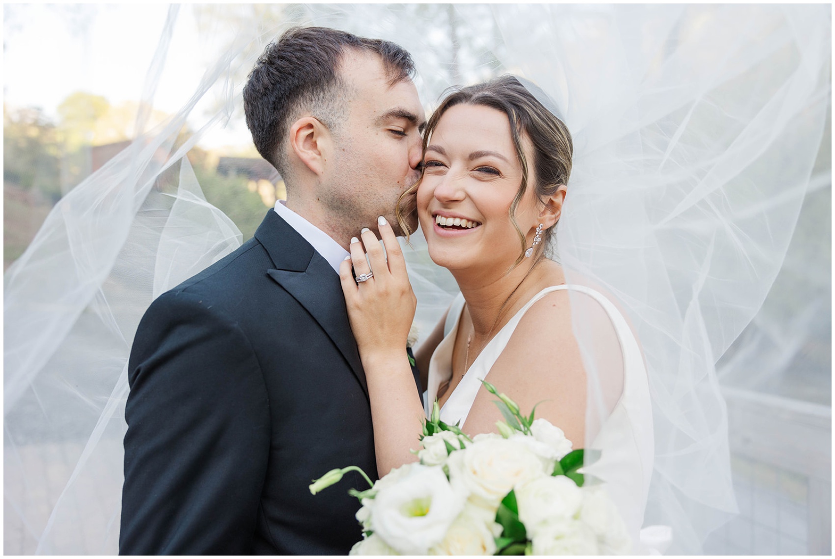A bride giggles while her groom kisses her cheek under the veil in the Roswell Mill Club Wedding venue gardens