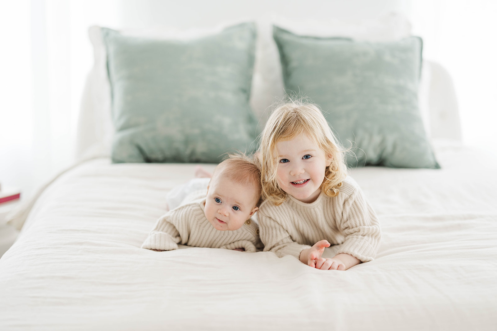 A toddler girl lays on a bed with her infant baby sibling in matching sweaters