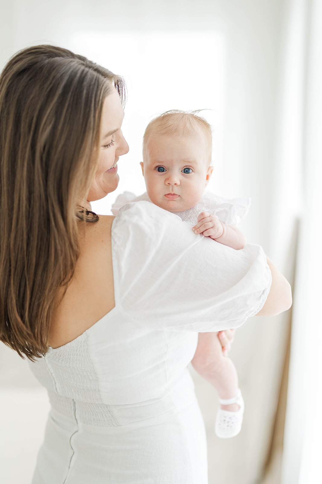 A happy new mom in a white dress holds her baby over her shoulder while standing in a studio after visiting baby boutiques in Atlanta
