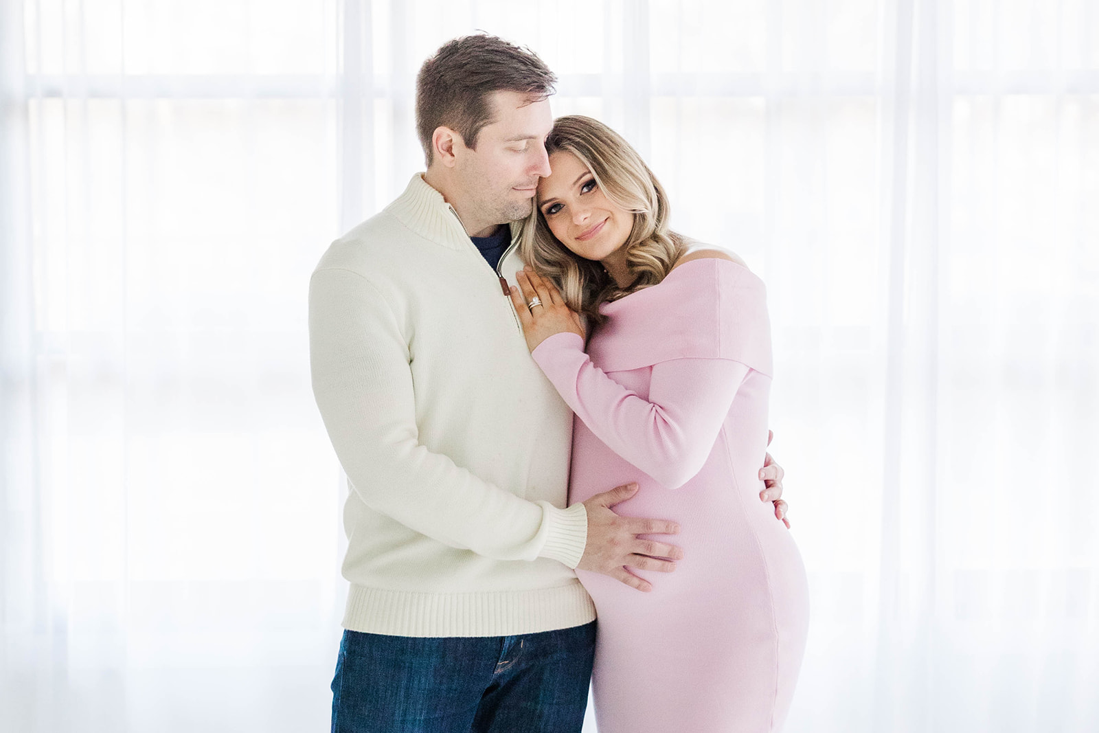 A happy pregnant couple snuggles while standing in large windows in a studio
