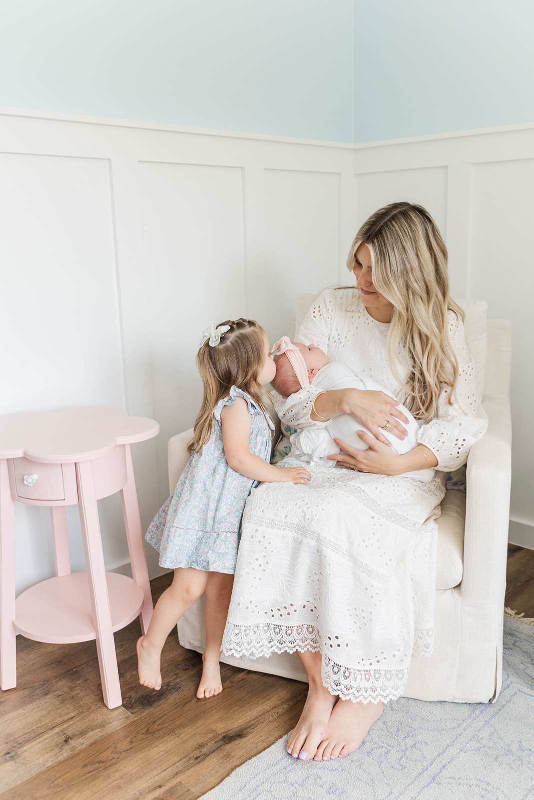 A toddler girl in a blue dress kisses her sleeping baby sister in mom's arms in a nursing chair