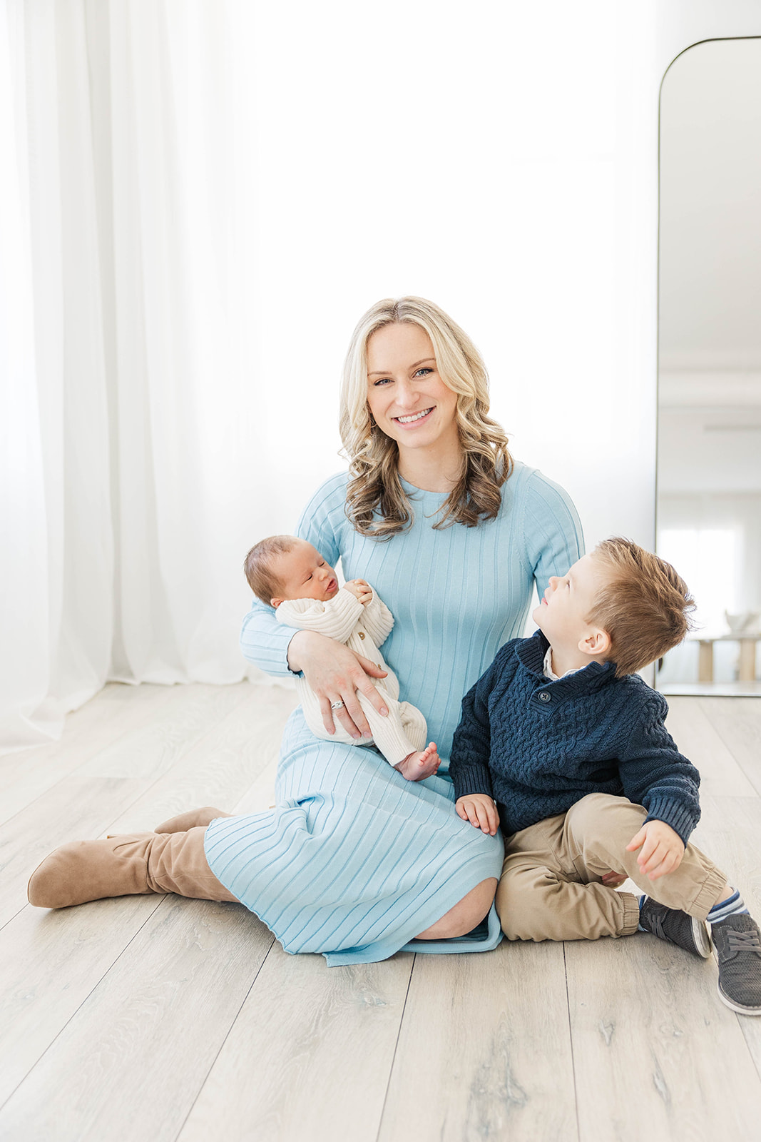 A mom smiles in a blue dress sitting on the floor with her newborn in one arm and toddler by her side after meeting Nannies in Atlanta