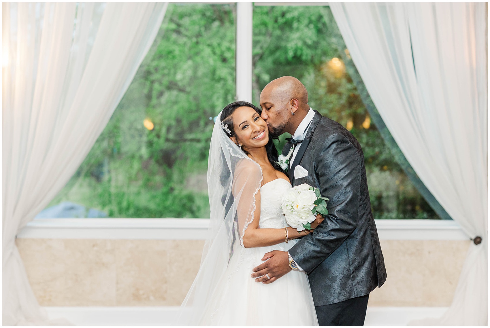 A groom kisses the cheek of his smiling bride in a window