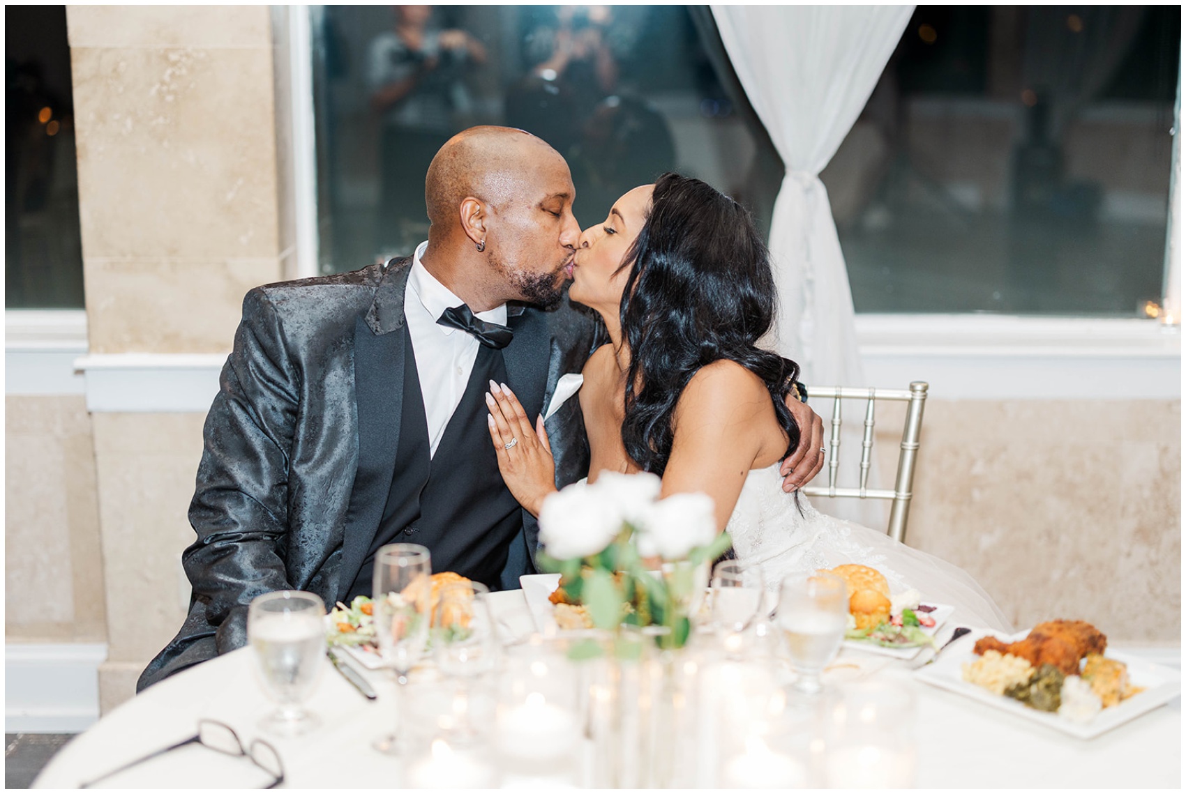NEwlyweds sit at their head table with plates of food and kiss