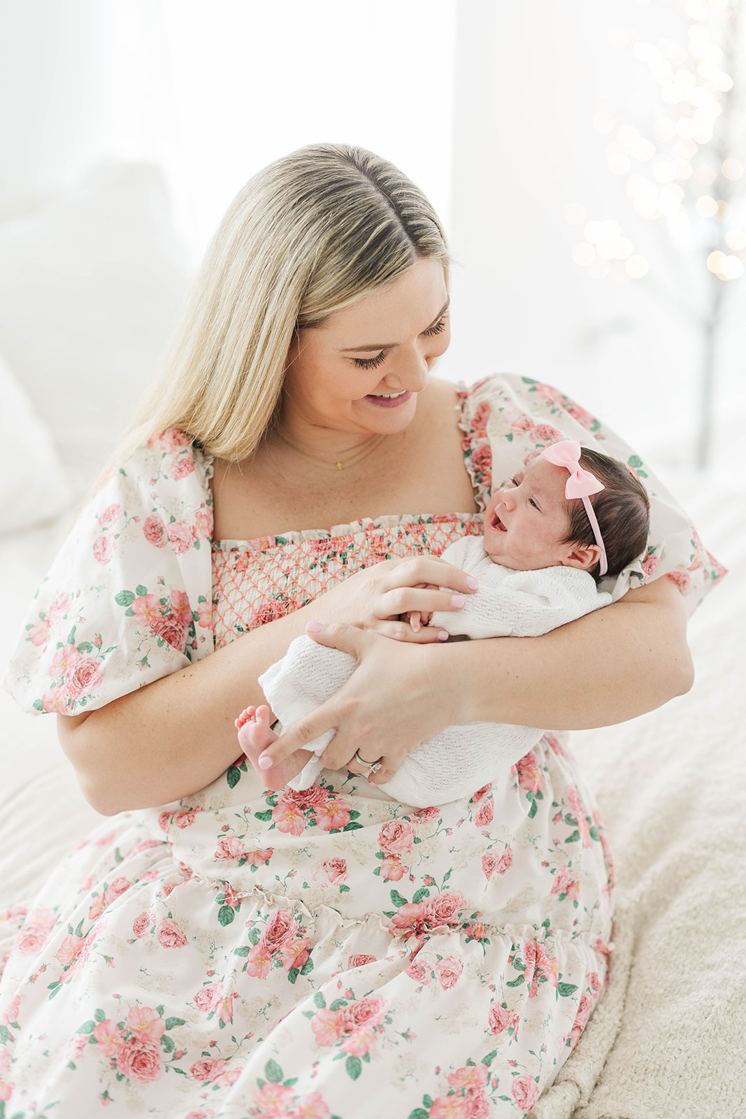 A happy new mom cradles her giggling newborn in a pink floral dress while sitting on a bed after meeting a postpartum doula in Atlanta