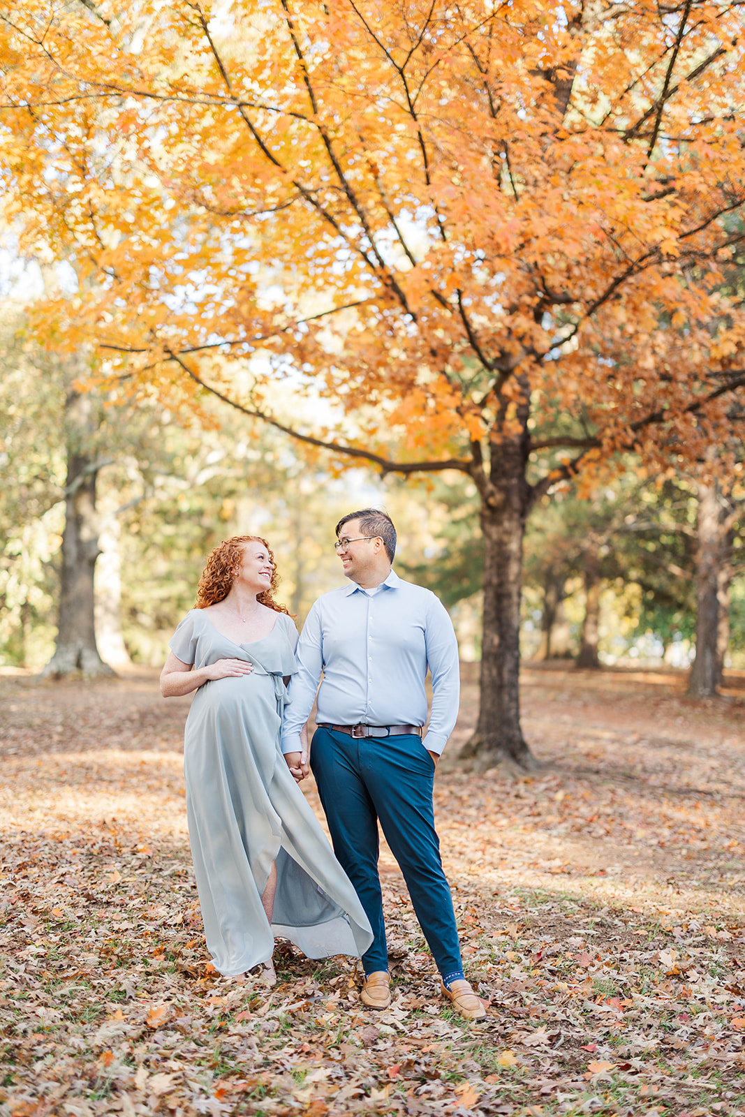 A pregnant couple holds hands while exploring a park in blue outfits in fall