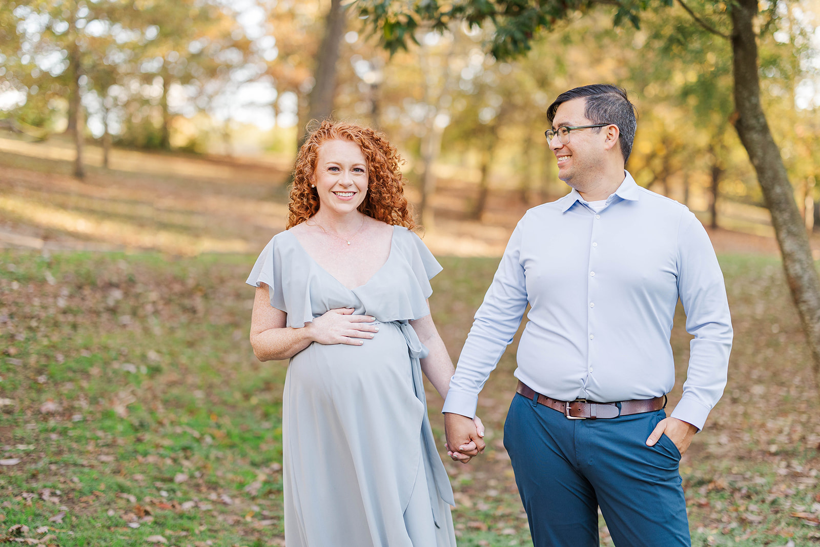 Happy expecting parents walk a park in fall smiling and holding hands in blue after a prenatal massage in Marietta, GA
