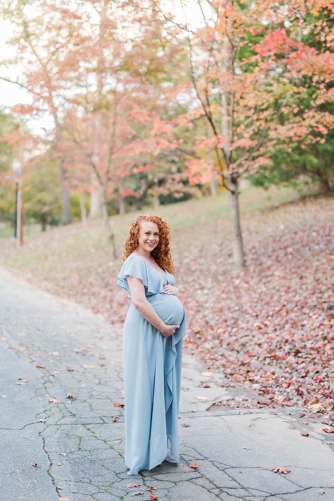 A smiling mother to be stands in a street in fall in a blue maternity gown after a prenatal massage in Marietta, GA