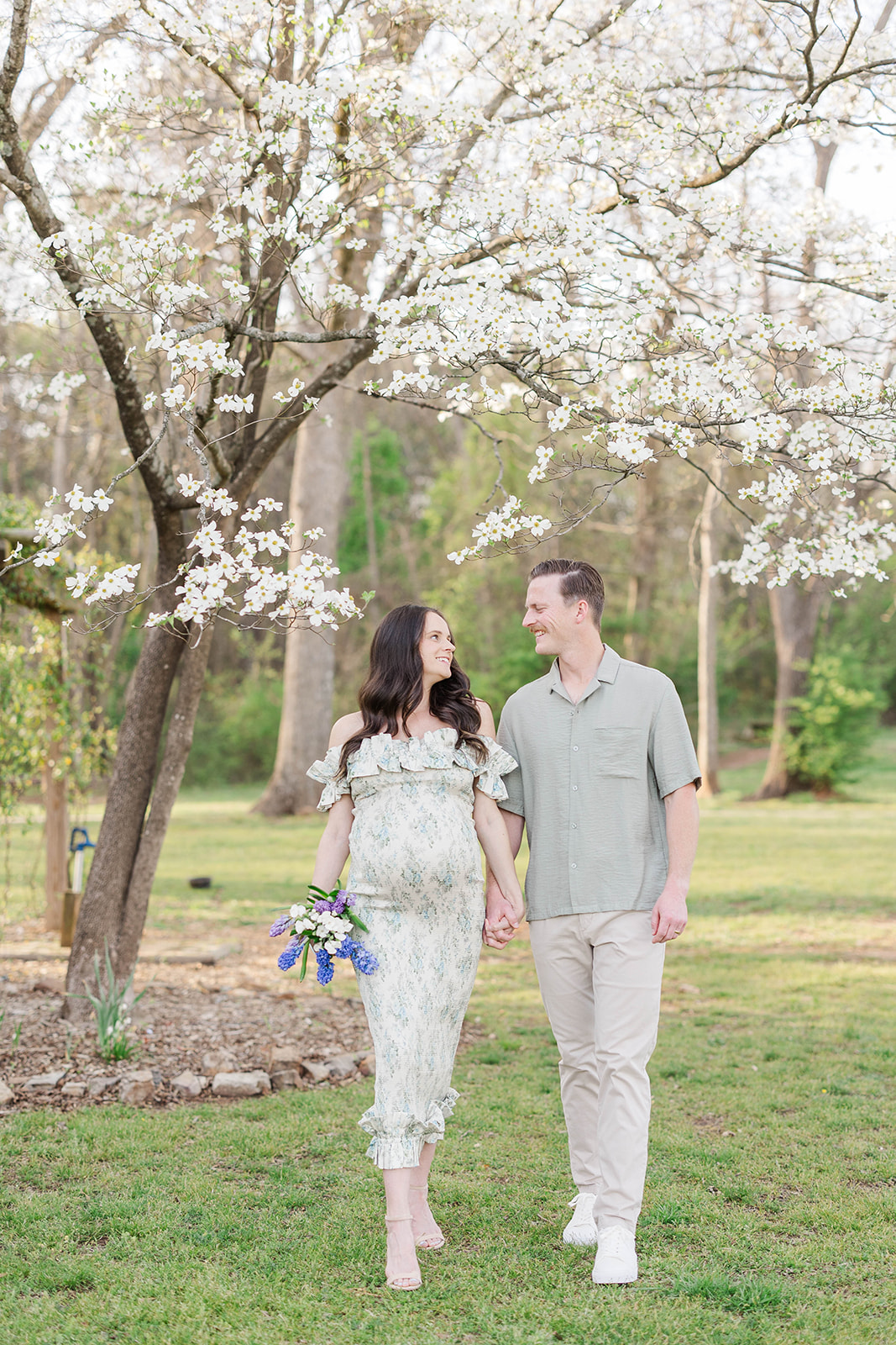 A happy pregnant couple walks under a blooming dogwood tree holding hands and smiling at each other