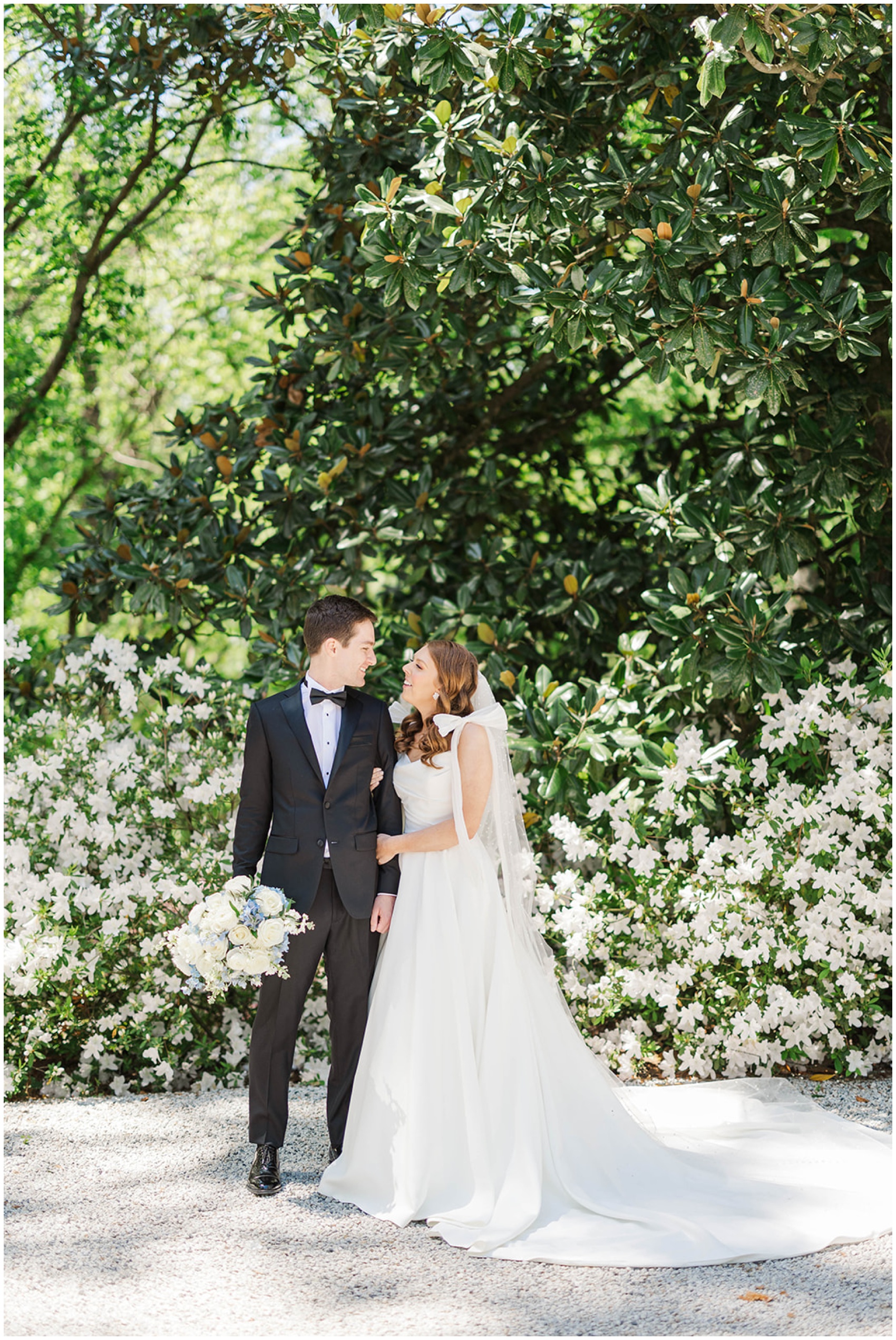 Newlyweds stand smiling at each other in front of white flowers