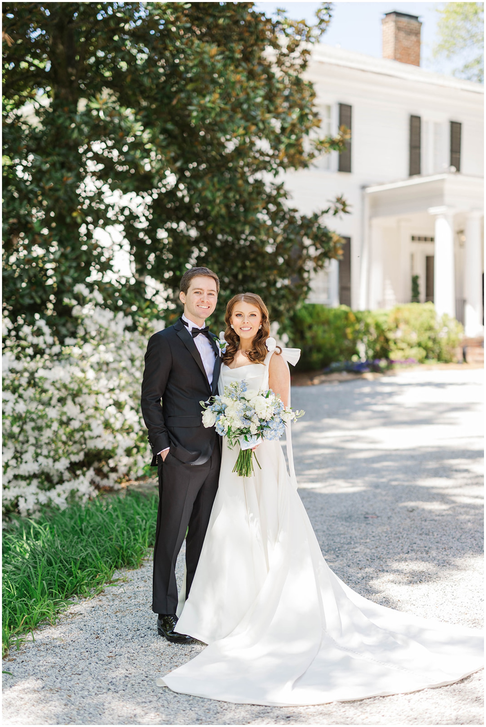 A bride and groom stand smiling together with a large white bouquet in the driveway
