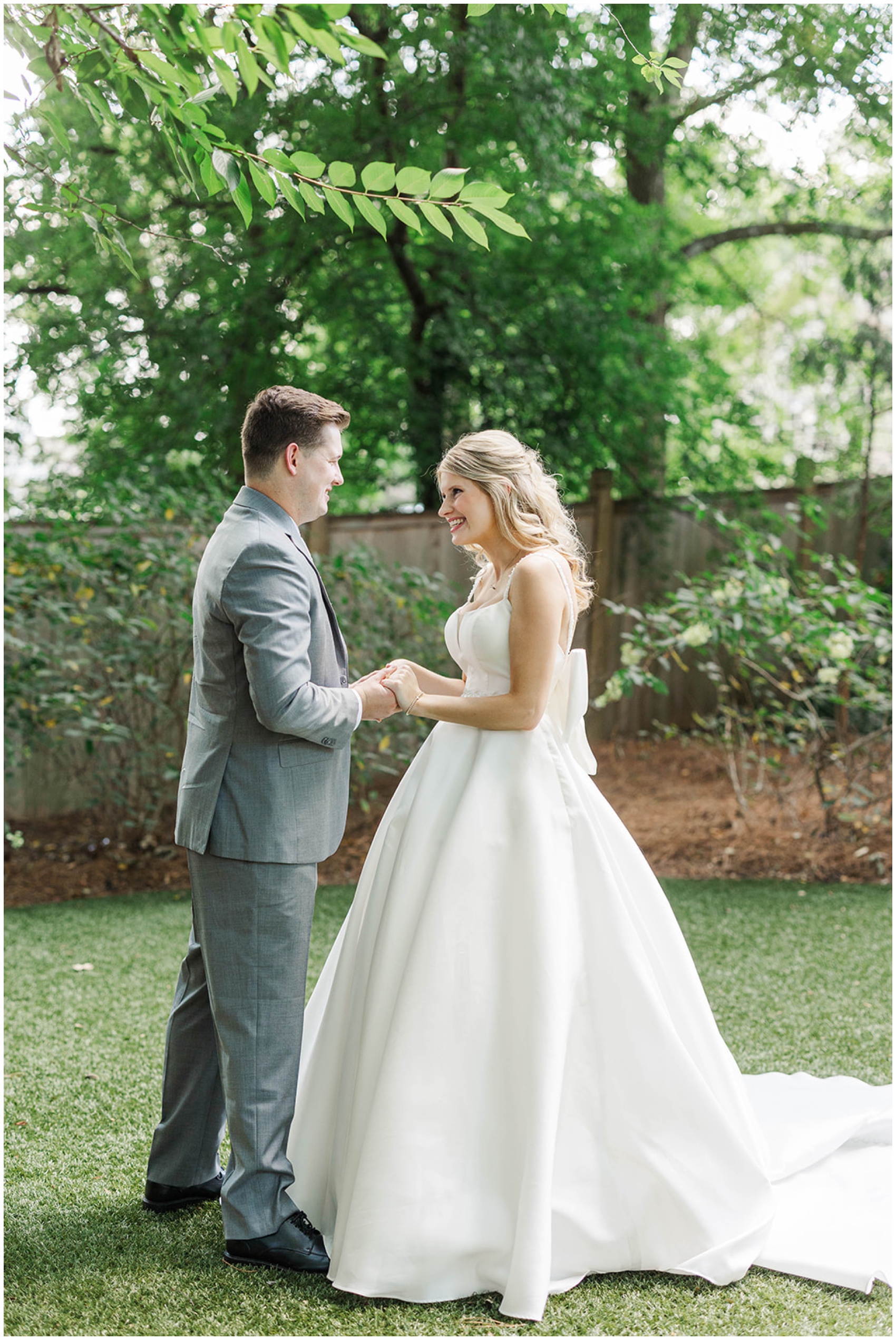 A bride and groom stand in a garden holding hands and smiling at each other