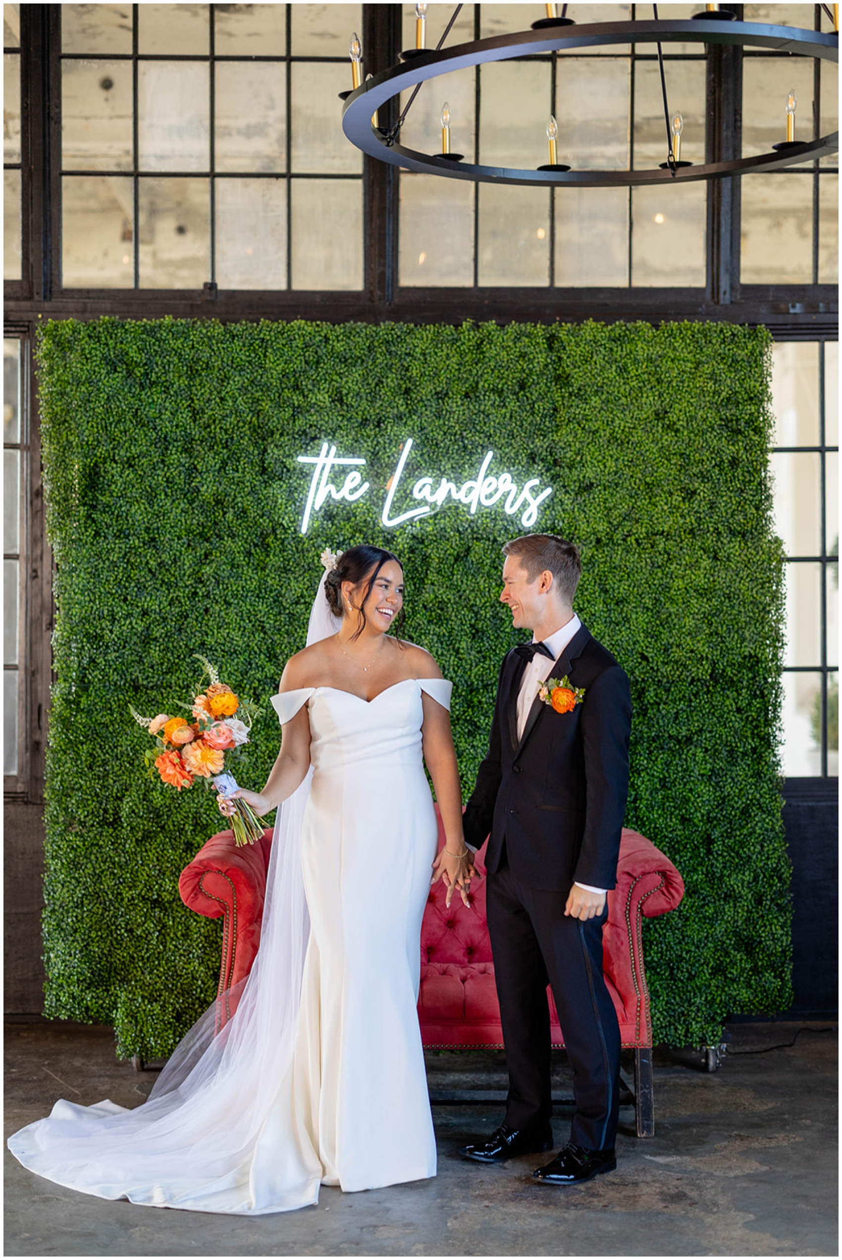 Newlyweds hold hands and laugh while standing in front of a custom neon sign on a plant wall