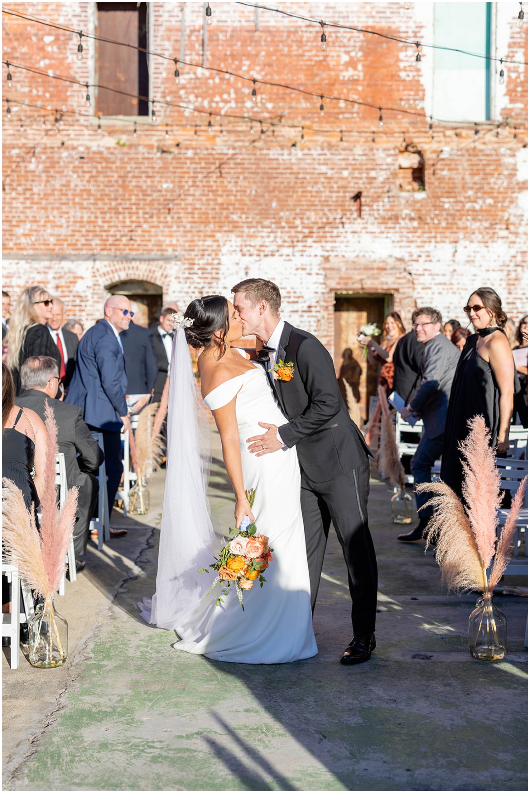 A groom kisses his bride while exiting their outdoor wedding ceremony as guests look on