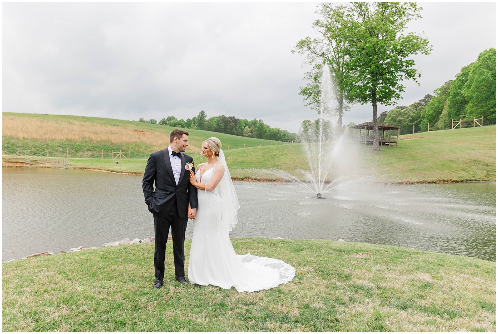 A bride snuggles on hr groom's arm while standing by the pond fountain during their Swan Lake Overlook Wedding
