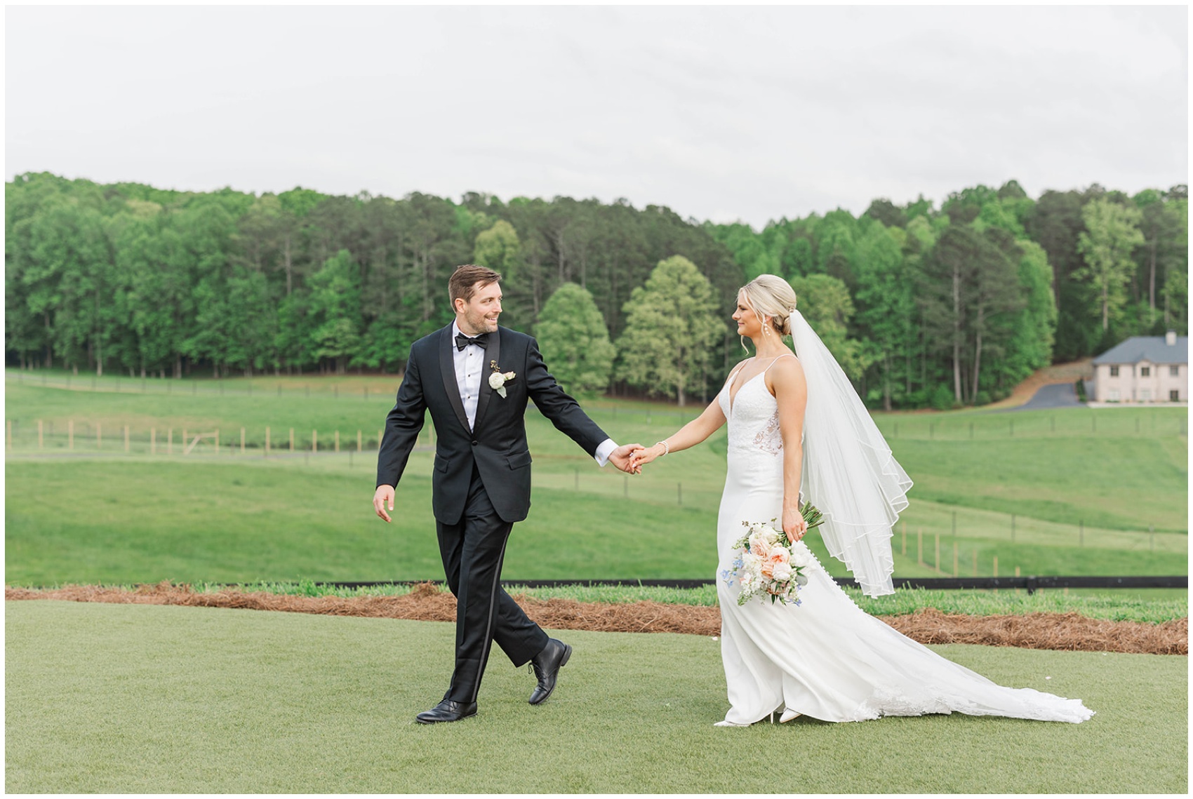 A groom leads his bride across a thick lawn by the hand as they smile at each other