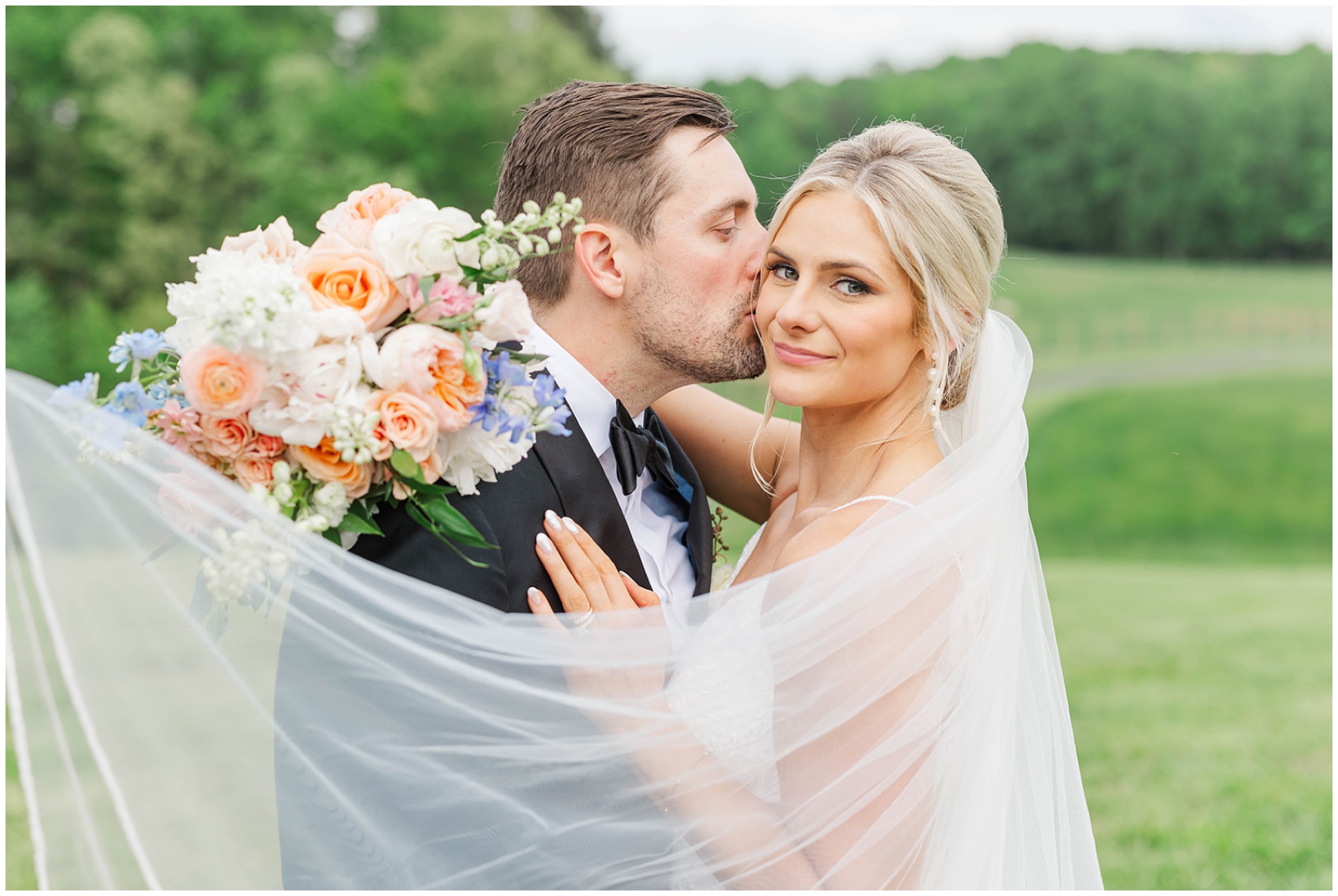 A bride smiles as her groom kisses her cheek in the hills while wrapped in the veil