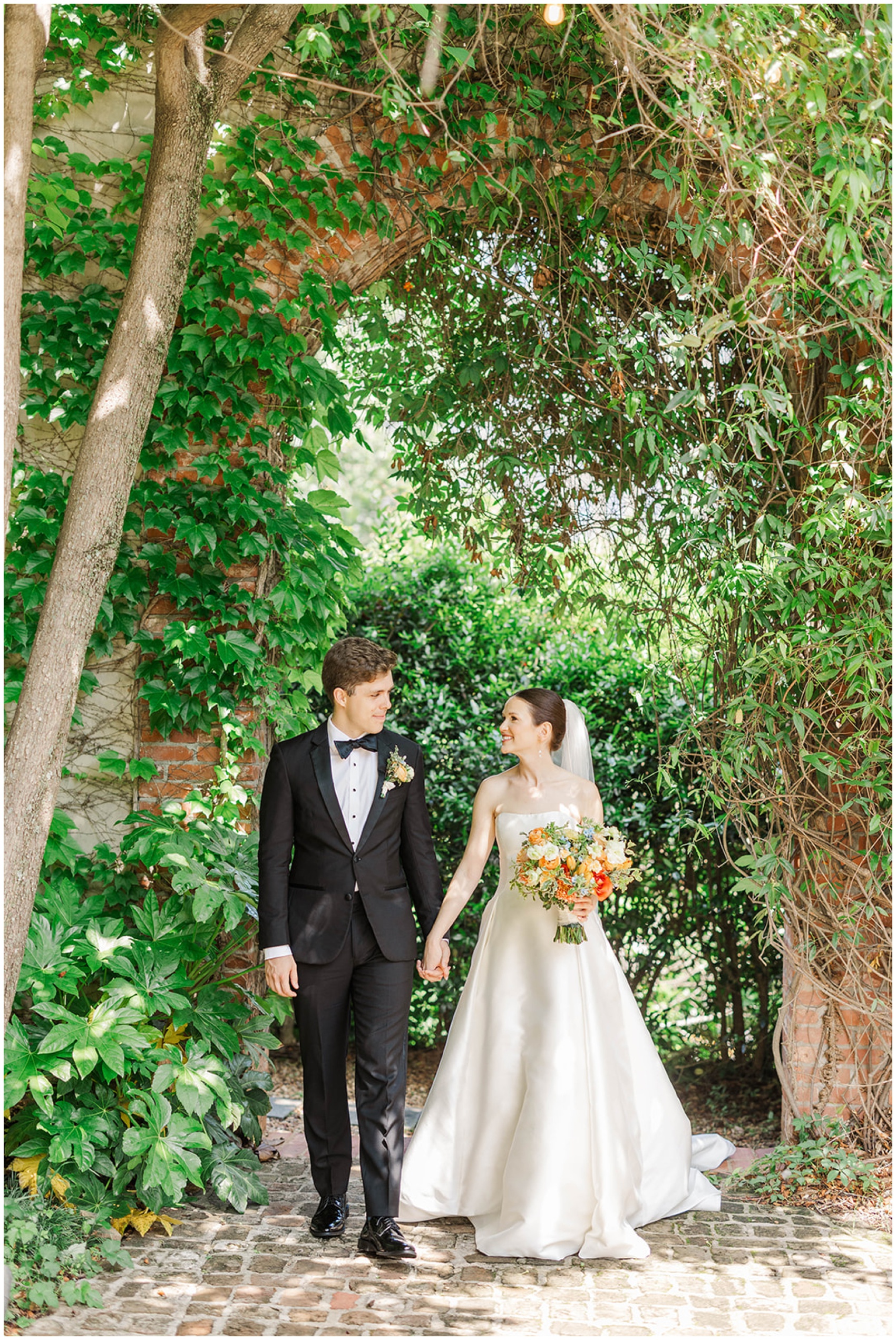 A bride and groom smile at each other while walking hand in hand under an ivy covered arch at their Summerour Studio wedding